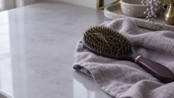 Hårbørste med tette børster og flere lange, løse hårstrå, lagt på en lilla linduk på et toalettbord i dagslys